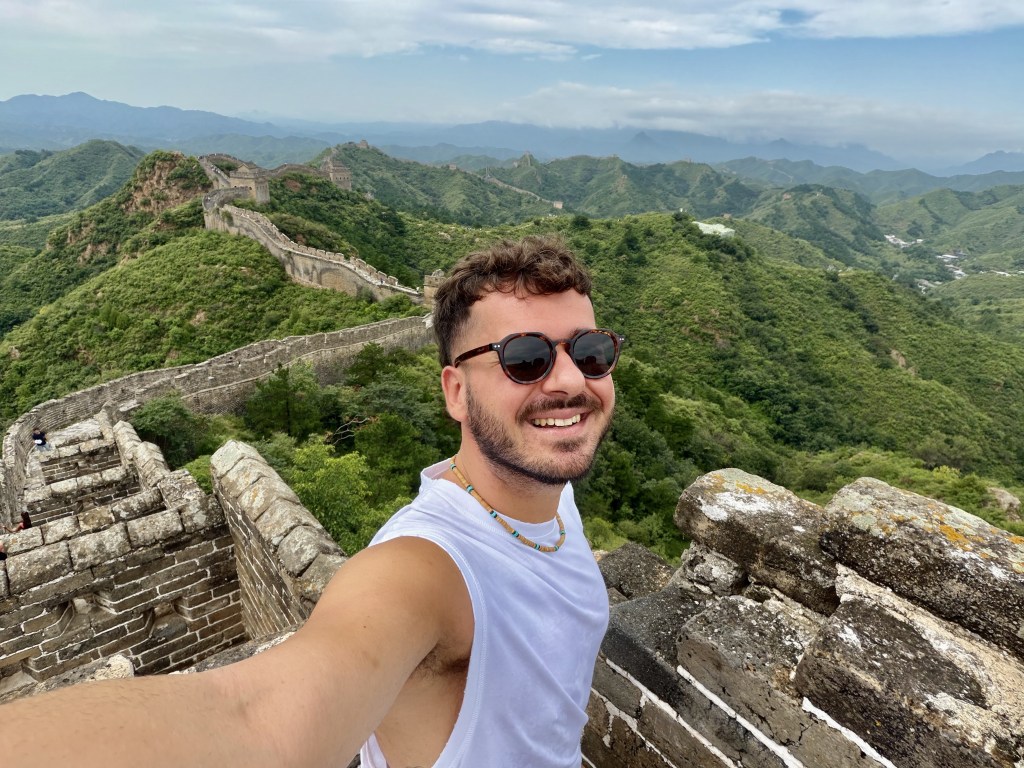 Un giovane in occhiali da sole sorride mentre scatta un selfie sulla Grande Muraglia Cinese, circondato da colline verdi e un cielo nuvoloso.